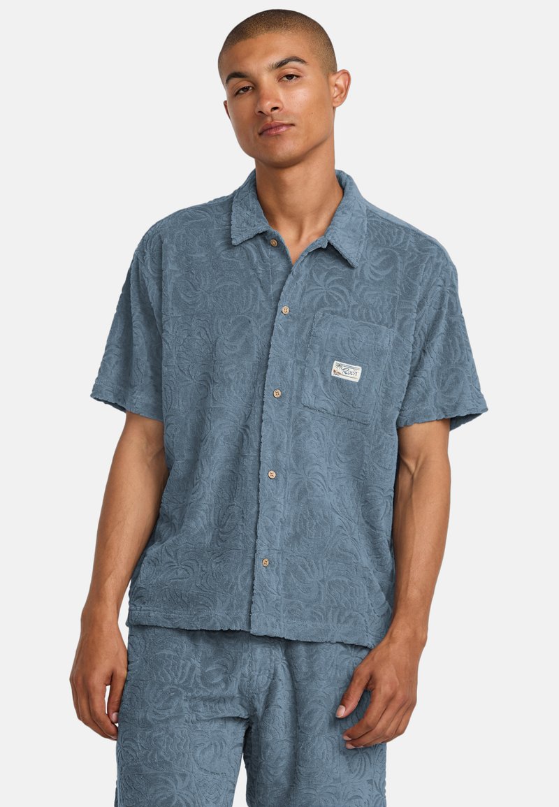 Camisa de botones de manga corta en tejido azul texturizado con un patrón floral y un bolsillo en el pecho con un parche de logo.