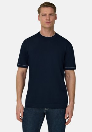 Uomo con capelli castano chiaro corti che indossa una maglietta a maniche corte blu scuro semplice e jeans blu scuro, in piedi contro uno sfondo bianco.