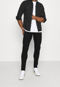 Camisa negra de botones, camiseta blanca debajo, jeans negros ajustados y zapatillas blancas con acentos negros. Diseño simple y moderno.