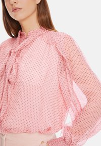 Rosa, durchsichtige Bluse mit Punkte-Muster, hochgeschlossenem Kragen, Rüschensleevs und einer Schleifen-Detail am Hals. Weiche Textur und leichtes Material.