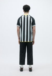 Svart og hvit stripegenser med korte ermer, gul hals, og svarte bukser. Skoene er svarte med røde detaljer; hvite sokker er synlige.