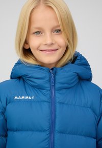 Blauer gepolsterter Mantel mit hohem Kragen und Kapuze. Verfügt über einen Reißverschluss auf der Vorderseite und das Logo "MAMMUT" in Weiß auf der linken Brust gestickt.