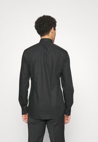 Man met krullend haar, gekleed in een zwarte blouse met lange mouwen en zwarte broek, staat tegenover een plain witte achtergrond.