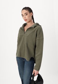 Vihreä fleece-pusero, jossa on puoli-vetoketju, huppu ja teksturoitu kangas. Ominaisuuksiin kuuluvat rento leikkaus ja roiskahtaneet olkapäät. Yhdistetty tummansinisiin farkkuihin.