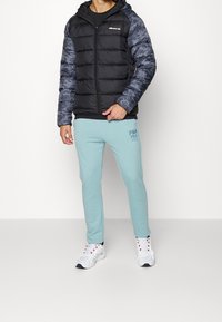  vadderad svart jacka med kamouflageblå ärmar, tillsammans med ljusblå Fila tracksuitbyxor och vita sneakers med röda accenter.