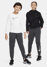 Twee kinderen dragen sportkleding: een wit longsleeve shirt met een zilveren logo en een zwarte hoodie met rits, gecombineerd met grijze sweatpants en zwarte sneakers.