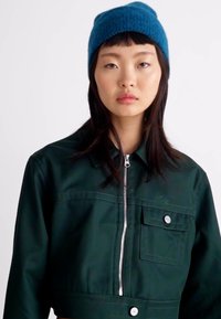 Chaqueta verde recortada con cierre de cremallera, detalle de bolsillo frontal y botones blancos; llevada con un gorro azul texturizado.