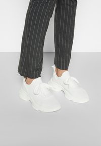 Hvide atletik sko med åndbart mesh-overdel, rund tå, flade snørebånd og kraftig sål, kombineret med mørke bukser med smalle striber.