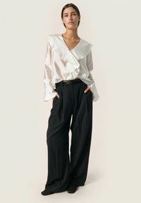 Blusa blanca con cuello de volantes y mangas largas, combinada con pantalones de raya diplomática de talle alto en color negro. Textura suave, diseño de pierna ancha.