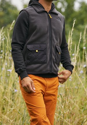 Mann im Freien, der eine schwarze Fleeceweste mit großen Vordertaschen und orangefarbene Hosen trägt, stehend zwischen hohem Gras und Wildblumen.