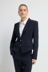 Granatowa marynarka na miarę z usztywnionymi ramionami, pojedynczym złotym guzikiem, klapami z wycięciem i dwoma przednimi kieszeniami, w połączeniu z białą koszulą na guziki.