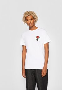 Hvit bomull t-skjorte med kort erm, med rød rose broderi på venstre bryst. Kombination med mørke jeans.