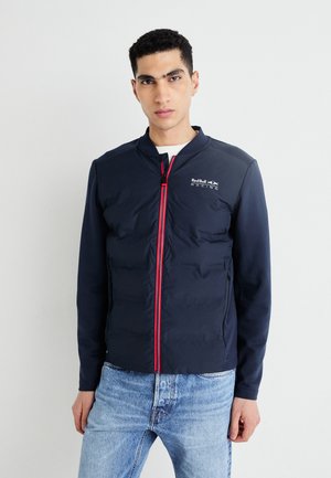 Marineblaue Steppjacke mit Stehkragen, Frontreißverschluss und rotem Akzent. Merkmale sind ein Logo auf der Brust und seitliche Taschen mit Reißverschluss. Strukturiertes Material.