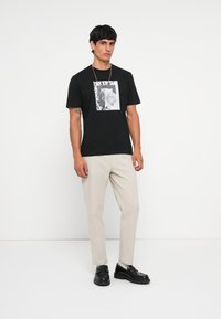 T-shirt noir avec un imprimé graphique, associé à un pantalon beige et des chaussures noires. Le mannequin se tient devant un fond blanc.