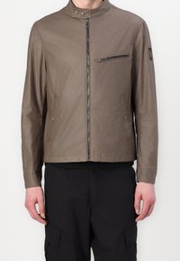 Olivově zelená, lehká bunda s vysokým límcem, předním zapínáním na zip a zipovanou náprsní kapsou. Vyznačuje se hladkou, jemnou strukturou a minimalistickým designem.