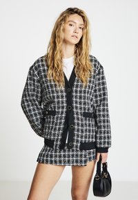 Black and white tweed jacket with large buttons, featuring side pockets and a black ribbed hem, paired with a matching skirt and small black bag.