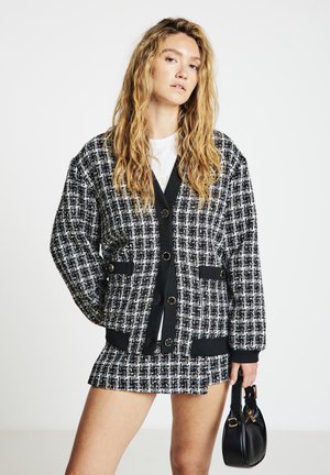 Schwarz-weißes Tweed-Jackett mit großen Knöpfen, seitlichen Taschen und einem schwarzen, gerippten Saum, kombiniert mit einem passenden Rock und einer kleinen schwarzen Tasche.