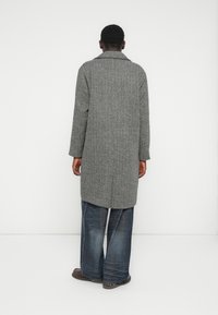 Pilkas žuvies kaulo rašto paltas su apykakle, ilgomis rankovėmis ir tiesiu kirpimu, derinamas su laisvais mėlynais džinsais ir tamsiais batais.