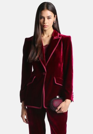Femme aux longs cheveux foncés portant un costume en velours rouge profond, tenant une petite pochette texturée assortie sur un fond clair uni.