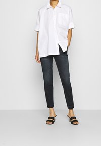 Valkoinen lyhythihainen kauluspaita, jossa on tasku, yhdistettynä tummiin kapeisiin farkkuihin ja mustiin remmisandaaleihin, esittelee rentoa ilmettä.