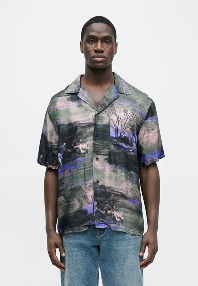 Νεαρός άνδρας που φοράει κοντομάνικο πουκάμισο με αφηρημένα μαύρα, μοβ και πράσινα σχέδια, συνδυασμένο με ανοιχτόχρωμο τζιν, στέκεται ακίνητος.