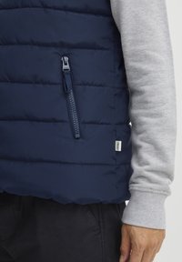 Tamsiai mėlynas pūkinis liemenės apšiltinimas su horizontaliais siūlais ir užtrauktuku užtrauktuku kairėje pusėje, dėvimas ant šviesiai pilkos ilgos rankovės marškinėlių.
