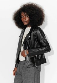 Veste de motard en cuir noir avec fermeture éclair asymétrique, accessoires en argent et poches latérales, portée sur un haut de couleur claire et un pantalon à motifs.