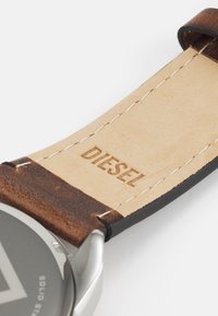 Bracelet de montre en cuir marron avec un revers beige, mettant en valeur des coutures blanches et le logo "DIESEL" embossé ; boîtier en métal argenté avec un cadran circulaire.