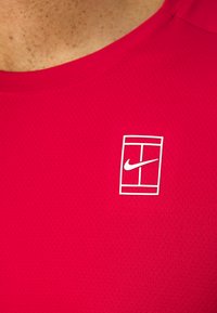 Rød idrettstrøye laget av pustende, teksturert stoff. Har et hvitt Nike-logo innenfor en rektangulær ramme på venstre bryst.