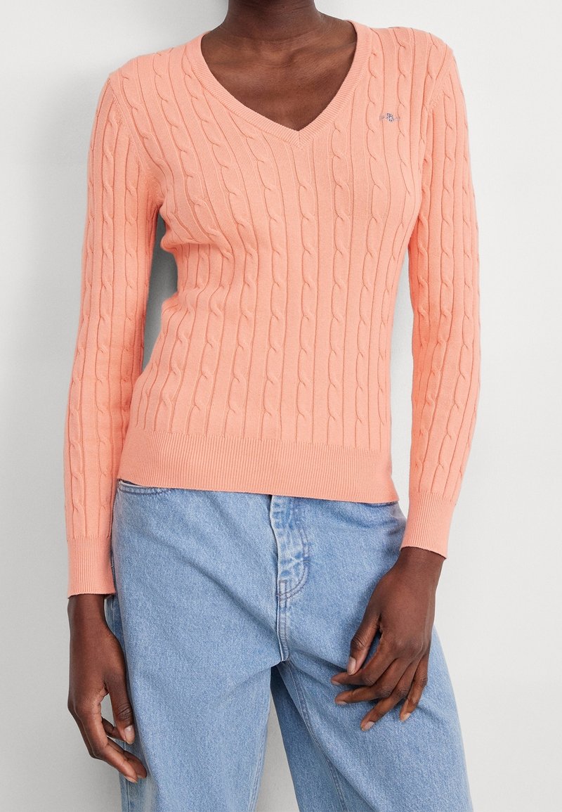 Personne portant un pull en maille torsadée couleur pêche à col en V avec manches longues et un jean taille haute bleu clair, sur un fond uni.