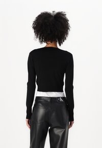 Schwarzer Pullover mit langen Ärmeln und Rundhalsausschnitt, kombiniert mit glänzenden schwarzen Kunstlederhosen, die ein Etikett am Taillenbund haben.