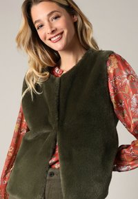 Olivgrüvester aus Kunstfell mit schlichter Schnittführung, ohne Verschlüsse. Tragen über einer roten Bluse mit floralen Mustern und langen Ärmeln. Glatte Textur.