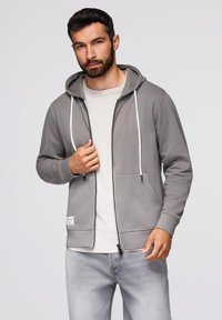 Szara bluza z zamkiem błyskawicznym i regulowanym kapturem, z przednimi kieszeniami oraz kontrastowym białym zamkiem. Nosi na sobie jasnoszary t-shirt i jasne dżinsy.