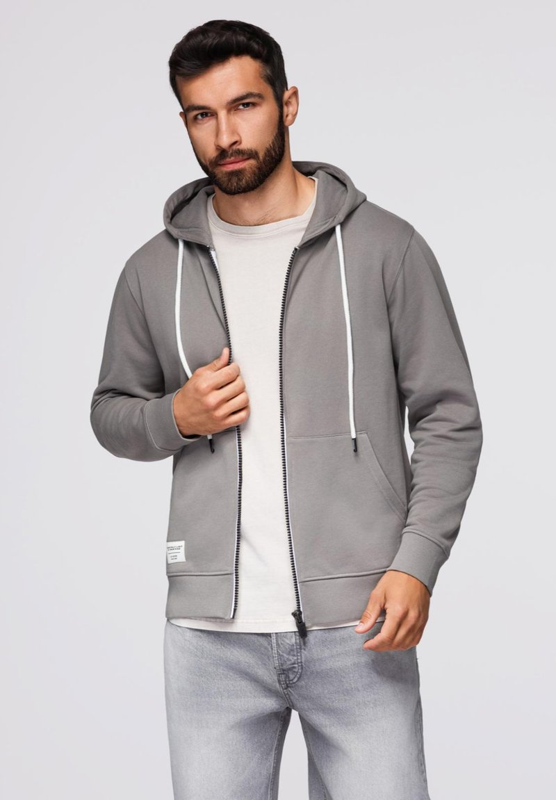 Szara bluza z zamkiem błyskawicznym i regulowanym kapturem, z przednimi kieszeniami oraz kontrastowym białym zamkiem. Nosi na sobie jasnoszary t-shirt i jasne dżinsy.