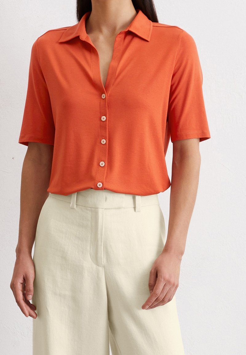 Femme portant une chemise orange à manches courtes boutonnée, rentrée dans un pantalon large crème taille haute, debout sur fond blanc.