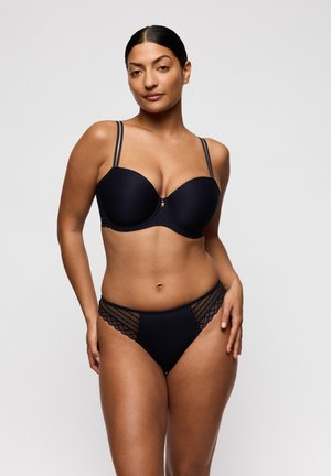 Vrouw met donker haar in een zwarte beha met dubbele bandjes en bijpassende slips met kantaccenten, staand tegen een effen lichte achtergrond.
