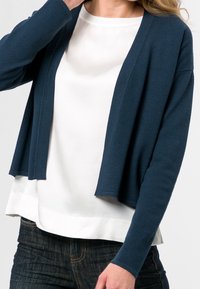Navy blue cardigan with textured pattern, open front, long sleeves, layered over a white top, paired with dark denim.