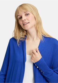 Světlá modrá cardigan vyrobená z měkkého materiálu, s uvolněným střihem, hlubokým výstřihem do V a knoflíkovými detaily na rukávech.