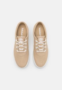 Beige canvas skor med vita snören, rund tå och texturerad ovandel. Har en logotyp på sidan och en vit gummisula.