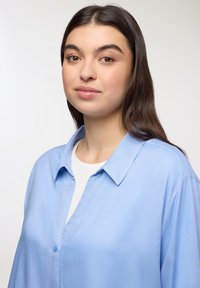 Jasnoniebieska koszula z guzikami, z kołnierzykiem, wykonana z gładkiego materiału, o luźnym kroju i widocznych guzikach z przodu.