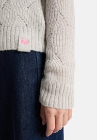 Helle graue Strickjacke mit geripptem Saum, die ein dezentes Wellenmuster aufweist und ein pinkes Herz-Logo an der Seite trägt. Kombiniert mit dunkler Jeans.