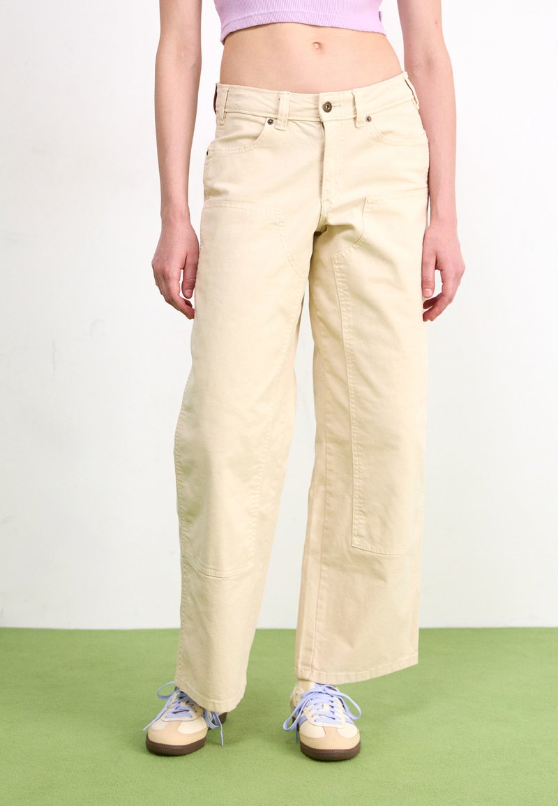 Pantalons larges de couleur beige clair en coton, dotés de deux poches avant et d'une taille haute. Associés à des baskets beige et bleues.
