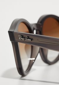 Harmaat muoviset aurinkolasit ruskeilla linsseillä, joissa on geometrinen kissankorva-muoto. Kehyksessä on hopeinen "Ray-Ban" -logo sivussa.