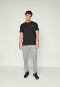 Giovane uomo in piedi che indossa una maglietta nera Puma, pantaloni sportivi grigi Puma e scarpe da ginnastica nere, con uno sfondo chiaro e semplice.