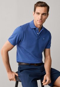 Homme portant un polo bleu et un short bleu marine assis sur un tabouret, regardant légèrement de côté sur un fond uni.