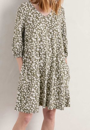 Kobieta w oliwkowozielonej sukience z dekoltem w serek, z białym kwiatowym wzorem i rękawami do łokcia, z rękami w kieszeniach, stojąca na tle gładkiej ściany.