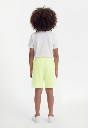 Niño con cabello rizado, vestido con camiseta blanca, pantalones cortos neon amarillos con bolsillos laterales y zapatillas blancas, de pie de espaldas sobre fondo blanco.