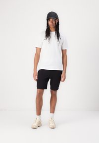 Camiseta blanca de cuello redondo, pantalones cortos negros y zapatillas beige con suelas texturizadas. La gorra es negra con visera curva. Un atuendo simple y casual.