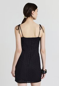 Eine Frau mit dunklem, zurückgebundenem Haar trägt ein ärmelloses schwarzes Minikleid aus Spitzenstoff mit dünnen Trägern, die zu Schleifen gebunden sind.