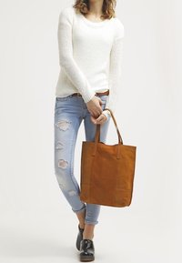 Hnědá kožená taška typu tote se dvěma dlouhými uchy, hladkou texturou, obdélníkovým tvarem a minimalistickým designem, nošená s lehkým svetrem a džínami.